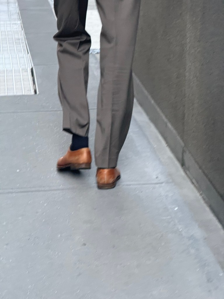 A person wearing gray trousers and brown shoes walking along a city sidewalk.