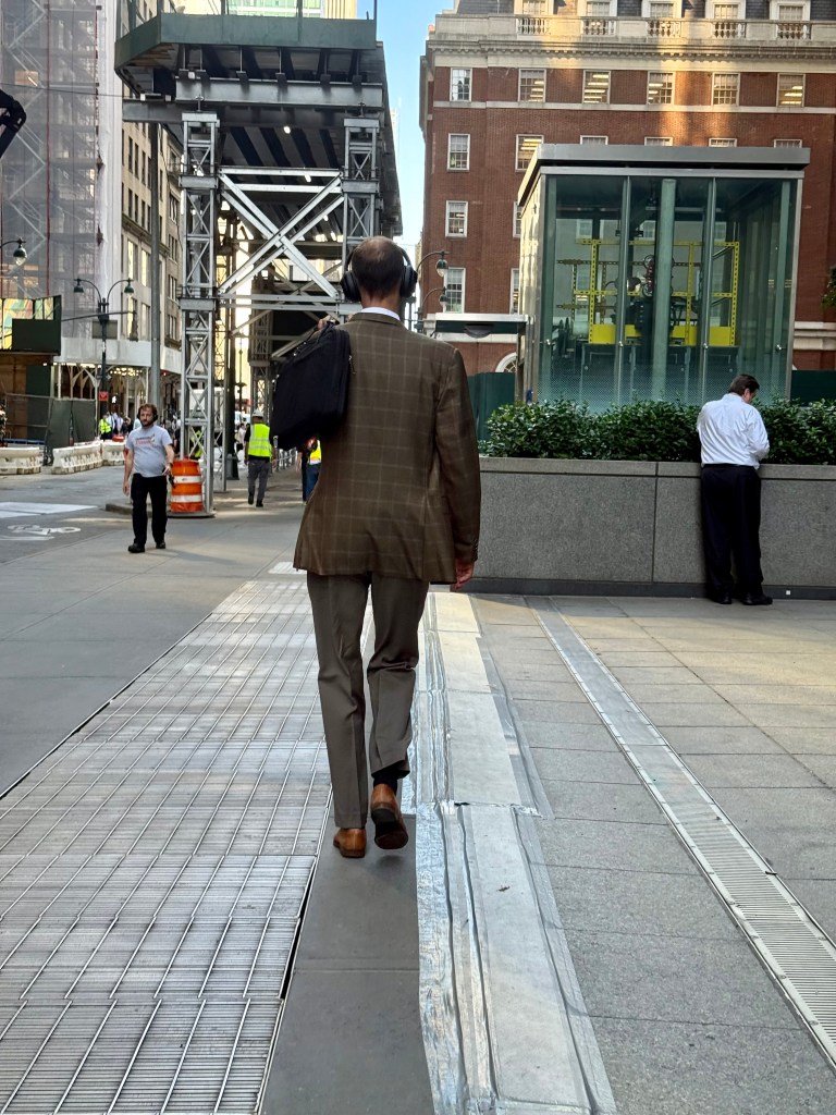 A man in a brown checkered suit carrying a black bag is walking down a city sidewalk, with construction and buildings visible in the background.