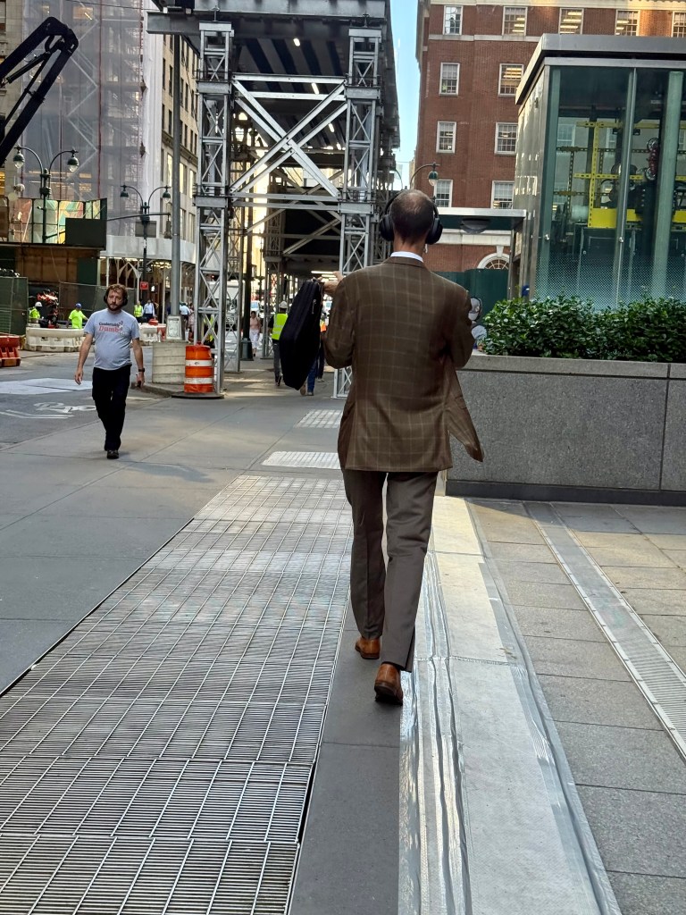 A man in a brown plaid suit and brown shoes walks away down a city street, wearing headphones and carrying a jacket. The background features construction scaffolding and other pedestrians.