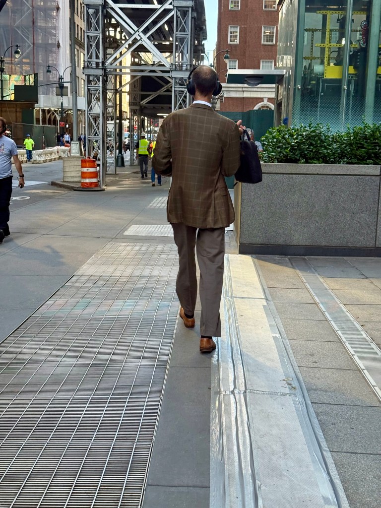 A man in a brown checkered suit and headphones walks down a city street, carrying a handbag. Construction scaffolding and people are seen in the background.