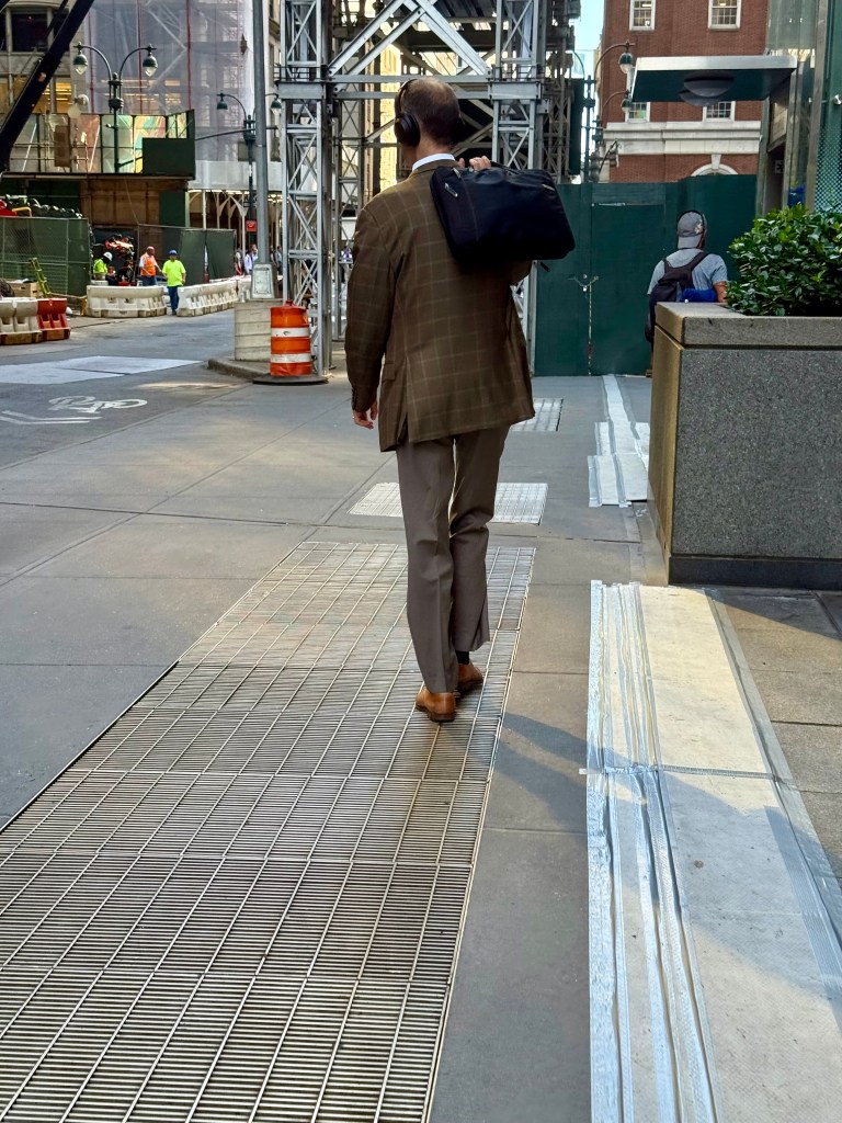 A man in a brown checkered blazer and beige slacks walks down a city sidewalk carrying a black bag. He is wearing headphones and heading towards a construction area.