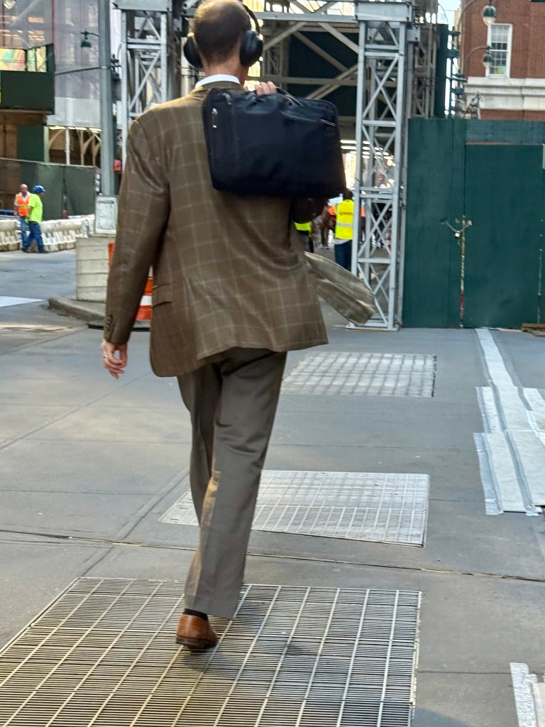 A person walking away, wearing a plaid blazer and carrying a black bag, with headphones on, in an urban environment.