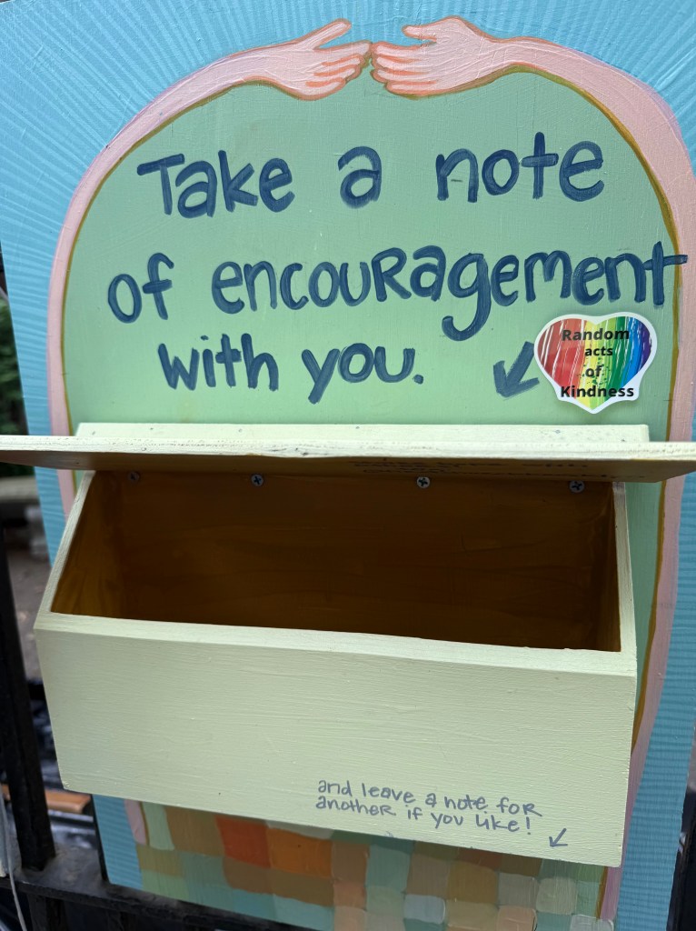 A colorful sign with two hands reaching towards each other, encouraging people to take a note of encouragement and leave a note for others in a designated box below.