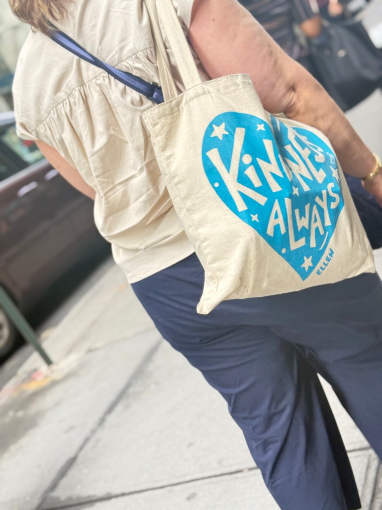 A person walking on the street with a beige tote bag that has a blue heart design and text that reads 'Kindness Always'.