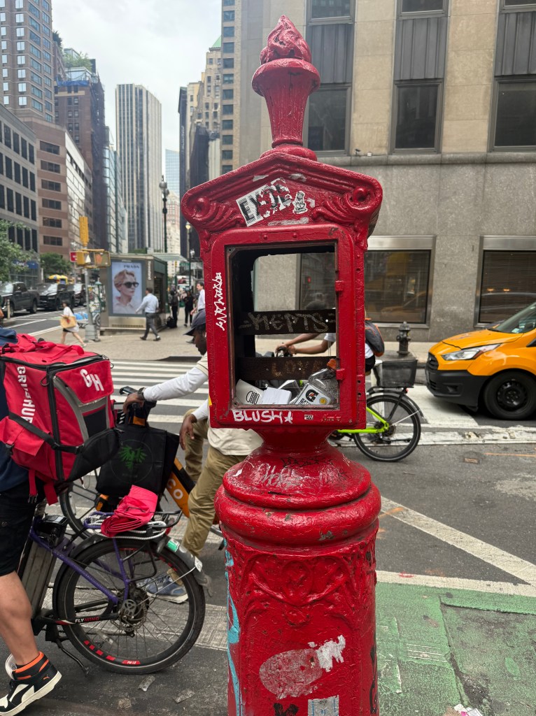 A red newsstand on a city street, covered in stickers, with people and bicycles in the background.