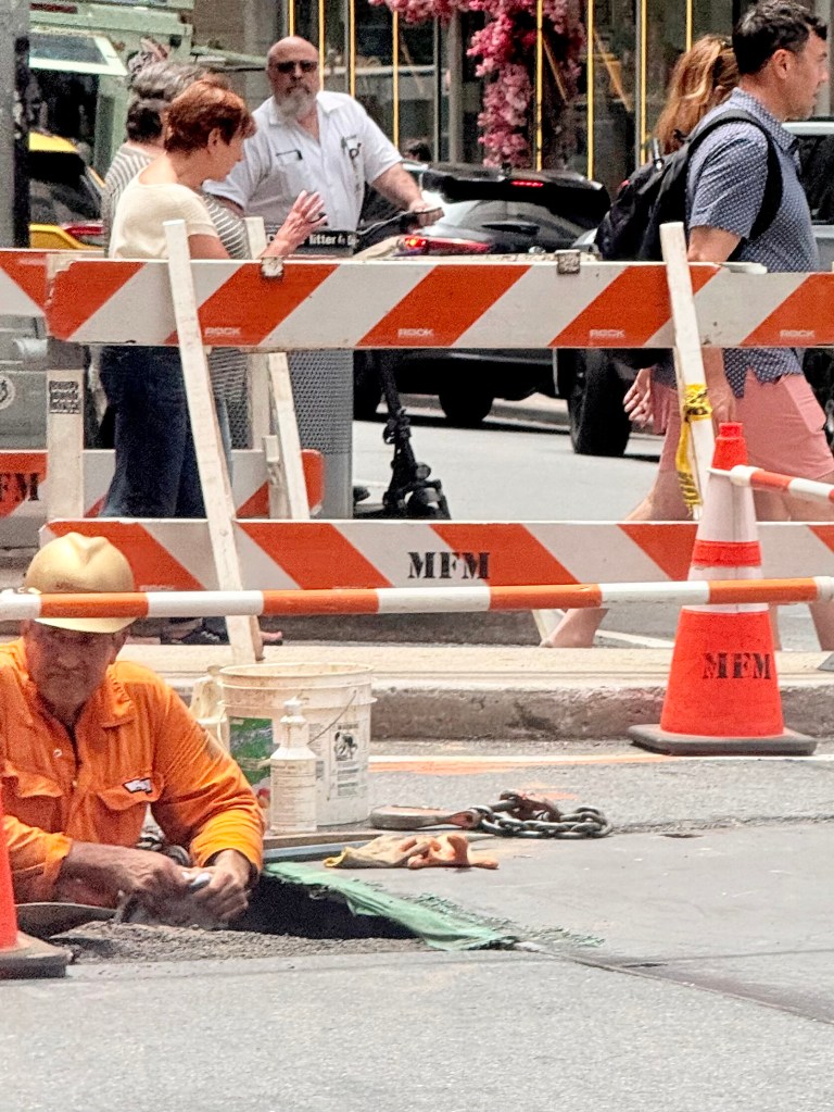 A construction worker wearing an orange shirt and hard hat is working on a street, surrounded by bright safety cones and barricades. Pedestrians are walking in the background, with flowering trees visible.