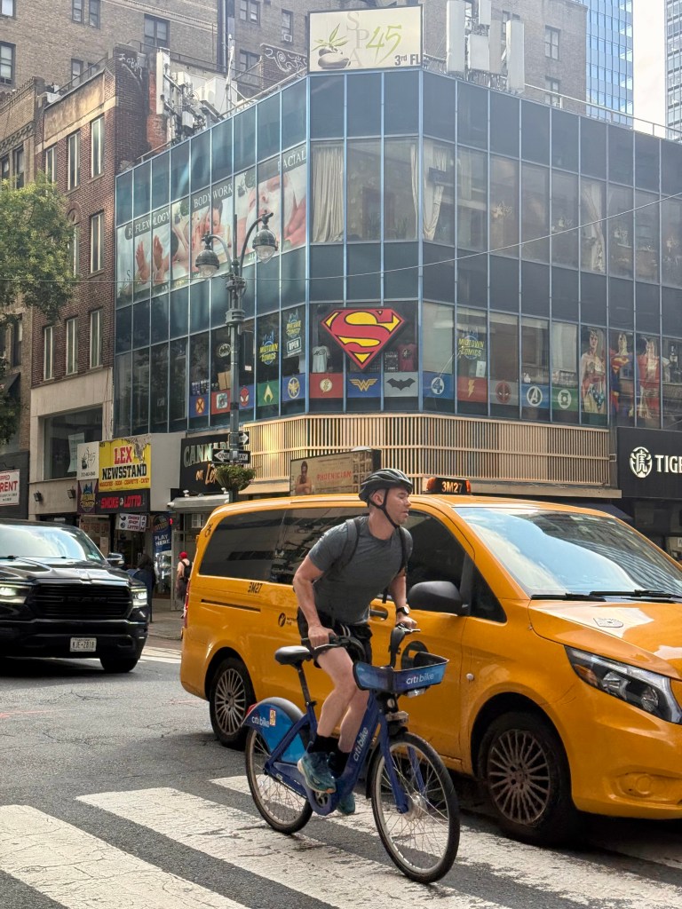 A cyclist riding a blue bike in a busy city intersection, with yellow taxis and a building featuring comic book hero logos like Superman and Wonder Woman.