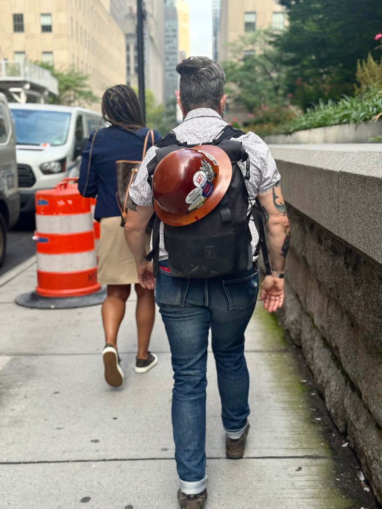 A person walking on a city sidewalk wearing a backpack and a construction helmet, with another person walking ahead, amidst urban surroundings and traffic.