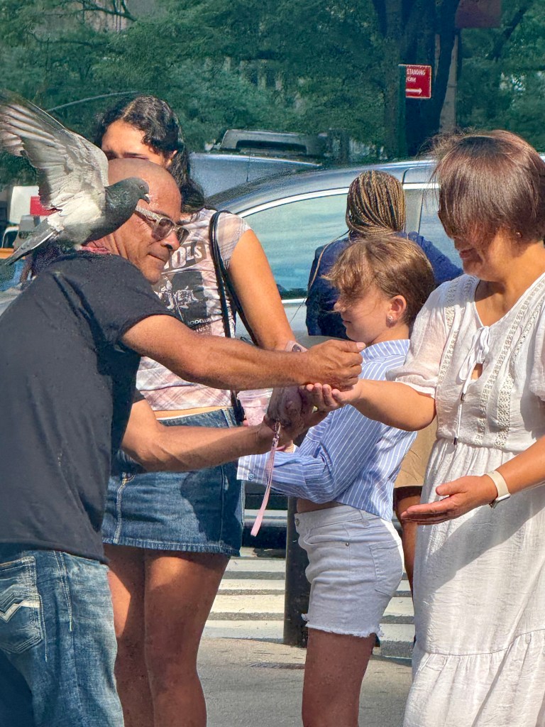 A man with glasses holds a pigeon on his arm while interacting with a group of people, including children, in a city street setting.