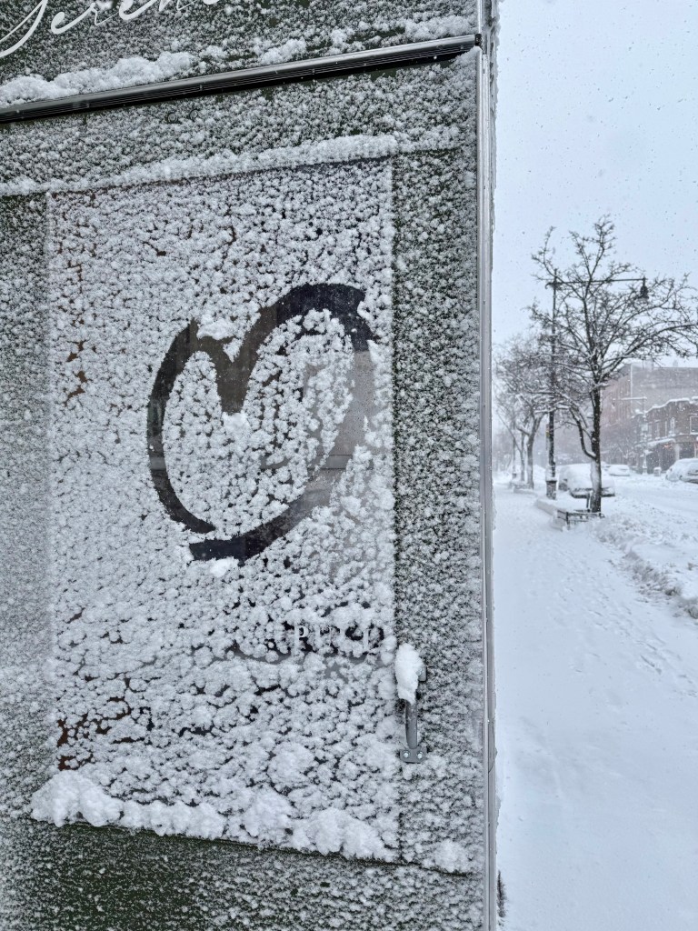 A snow-covered surface with frost, featuring a heart shape drawn in the frost. The background shows a snowy street with trees and benches.