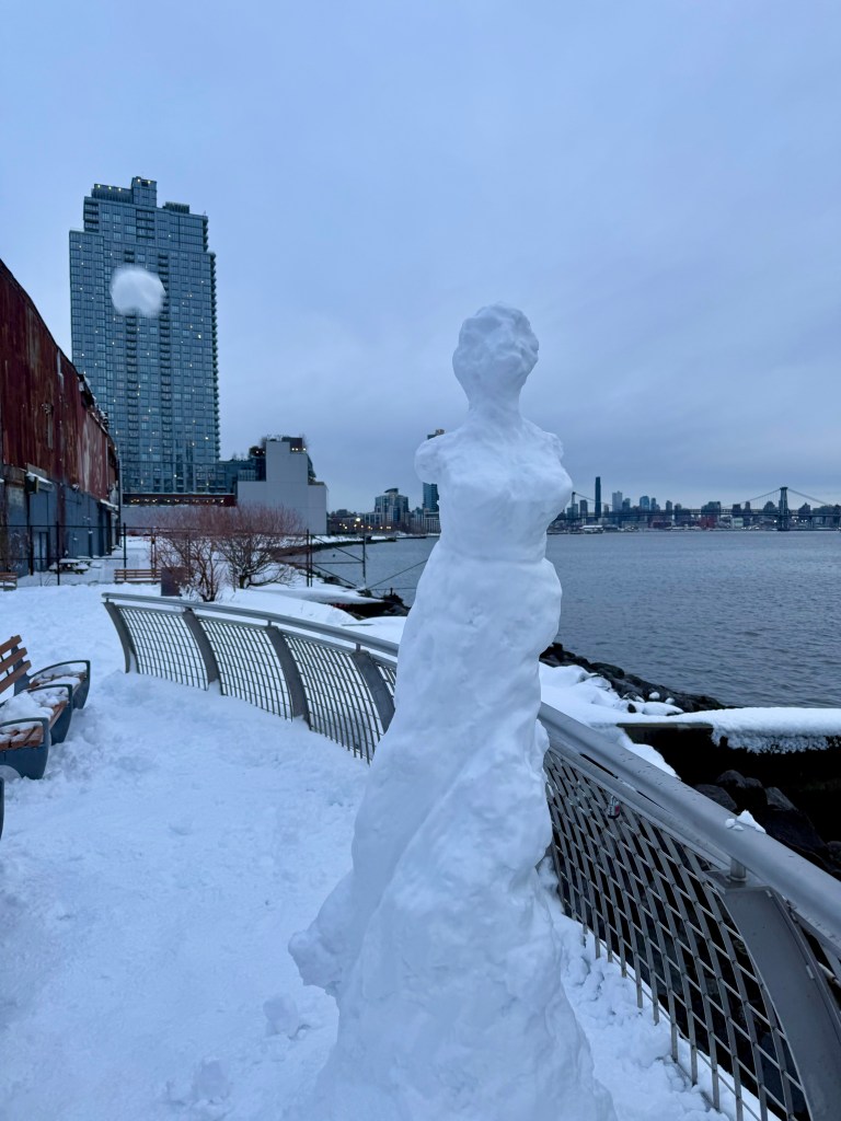 A snow sculpture resembling a figure stands on a snowy waterfront pathway, with a city skyline in the background under overcast skies.