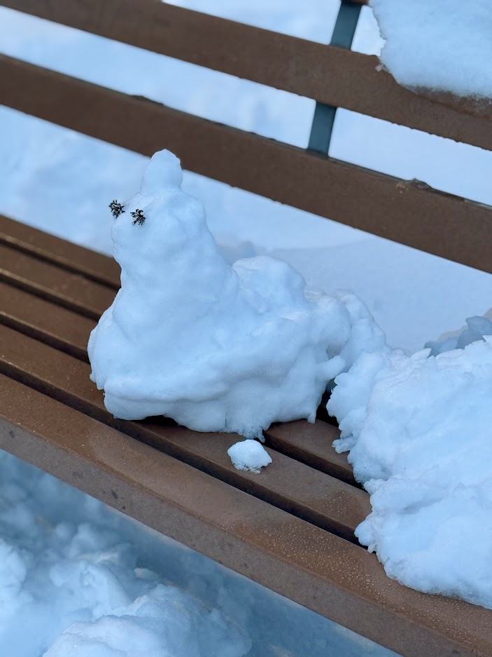 A snow sculpture resembling a small figure sits on a wooden park bench, with a snowy background.