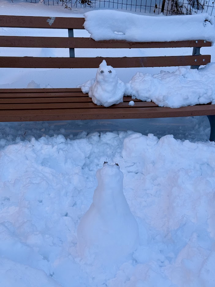 A snowman sitting on a bench, with a smaller snow figure in front of it, surrounded by freshly fallen snow.