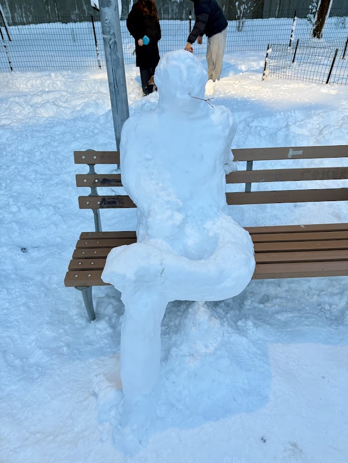 A snow sculpture resembling a person sitting on a bench, surrounded by a snowy landscape.