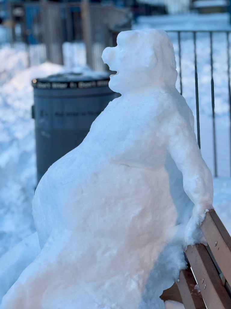 A snow sculpture resembling a sitting figure with a rounded belly, positioned on a bench, in a winter setting.