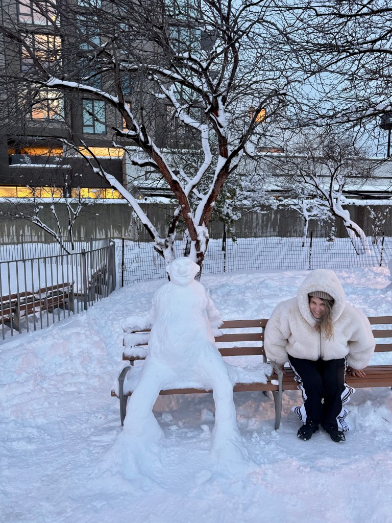 A woman in a fluffy white coat sitting next to a snow statue that resembles a person, on a snowy bench with a winter landscape in the background.