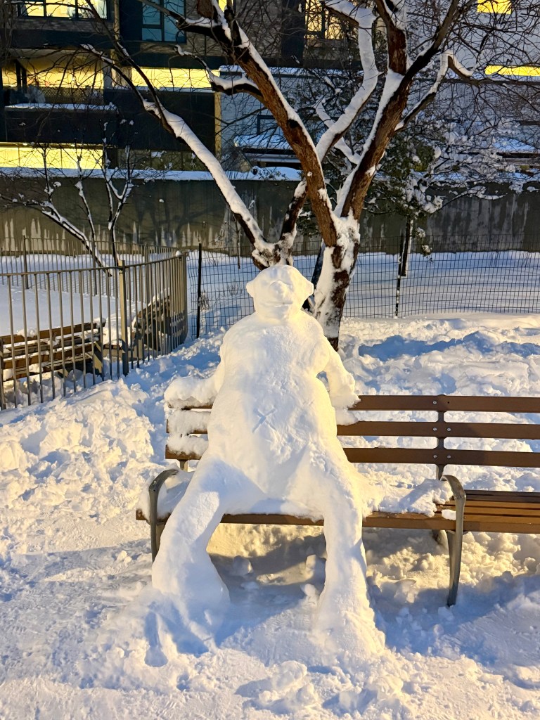 A snow figure resembling a person sitting on a park bench, surrounded by a snowy landscape and a tree in the background.