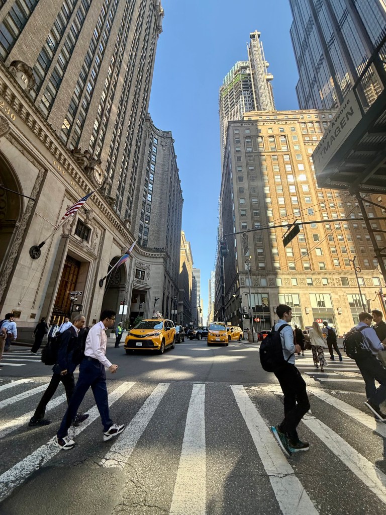 Busy street scene in a city with tall buildings, pedestrians crossing at a crosswalk, and yellow taxis on the road under a clear blue sky.