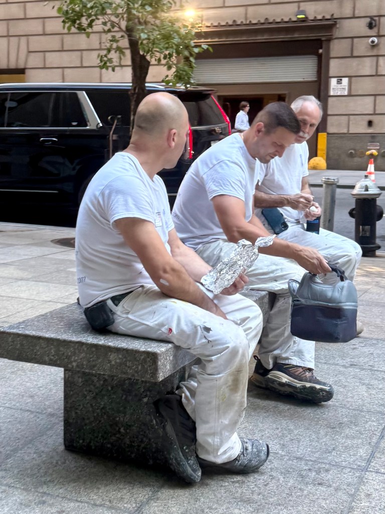 Three men sitting on a stone bench, wearing white shirts and paint-stained pants, enjoying lunch in a city street.
