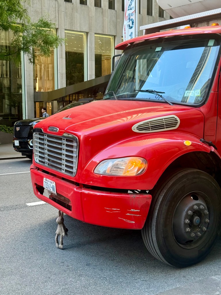 A red truck parked on the street with a small stuffed animal hanging from the front bumper.