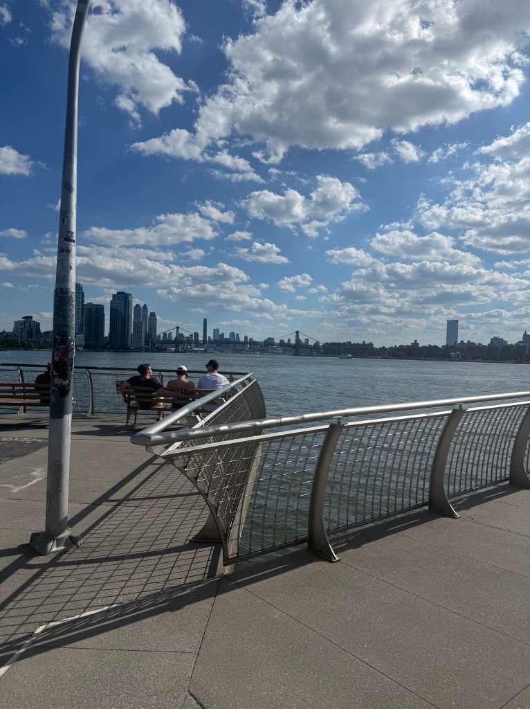View of a waterfront promenade with two people sitting on benches, surrounded by a city skyline and blue sky with clouds.