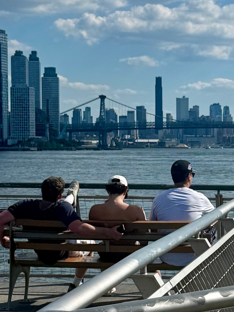 Three people sitting on a park bench by the waterfront, with a view of a city skyline and a bridge in the background.
