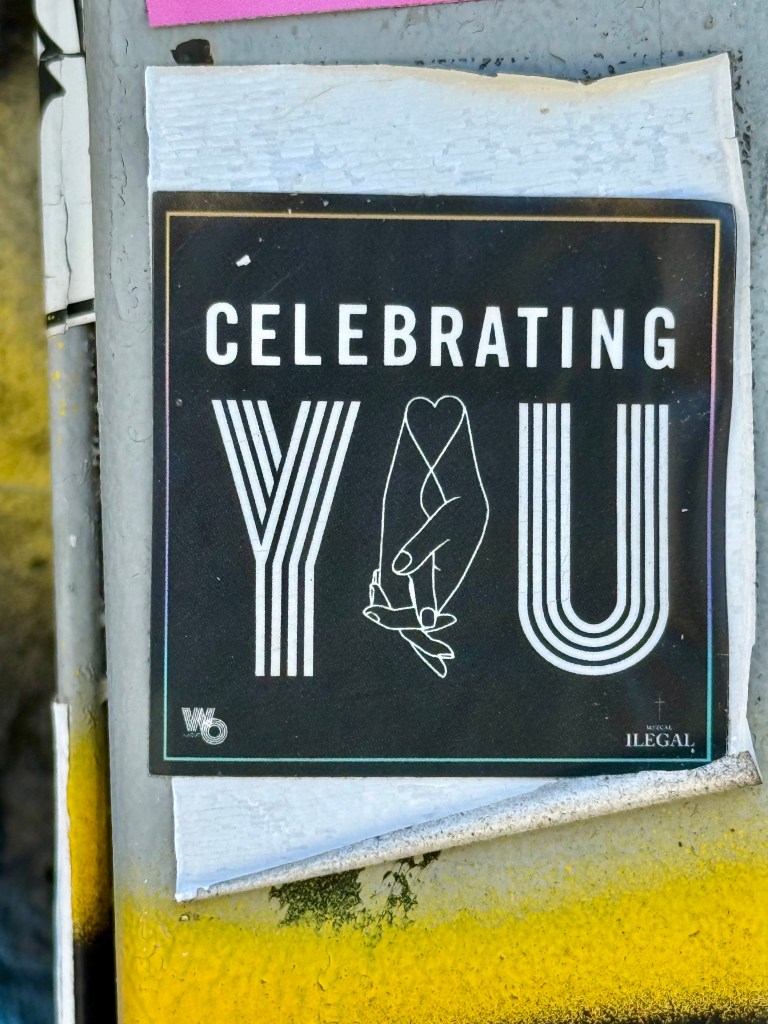 A sticker with the text 'CELEBRATING YOU' in bold white letters, featuring a graphic of a hand holding a small figure, set against a black background.
