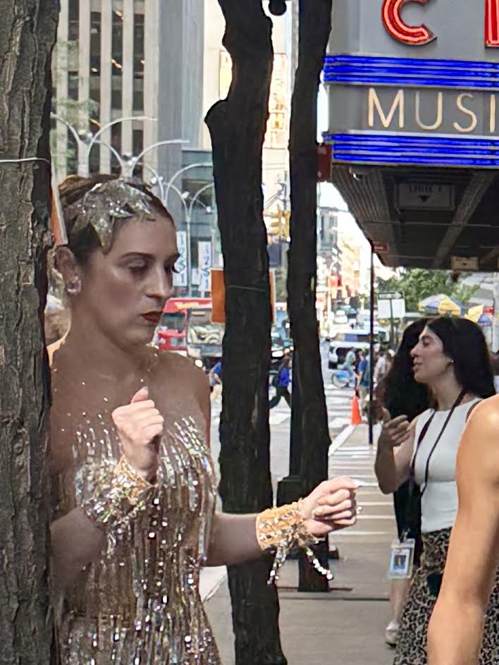 A woman wearing a sparkling gold dress and accessories stands next to a tree on a busy street, looking contemplative. In the background, another woman interacts with a friend, with city buildings and a theater marquee visible.