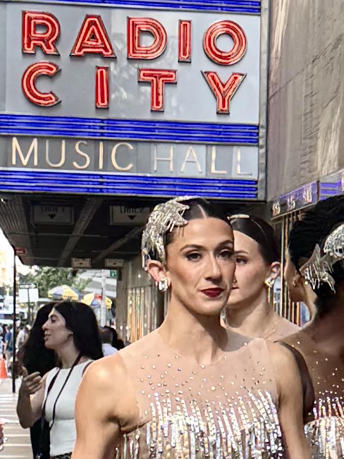A group of dancers dressed in sparkling costumes walking in front of the Radio City Music Hall sign in New York City.