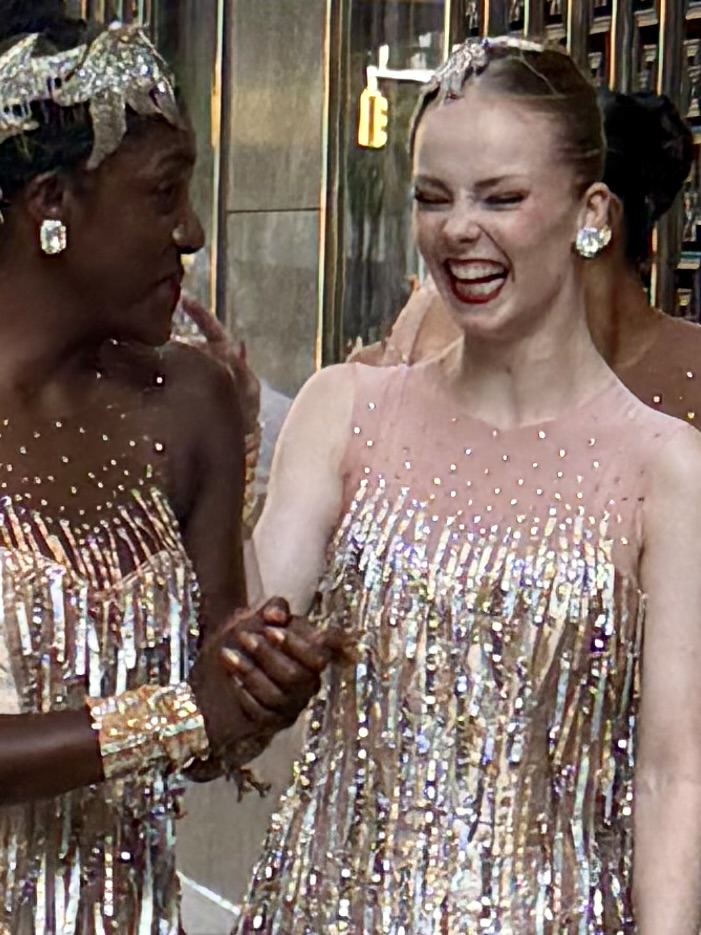 Two women in glamorous, sequined outfits smiling and holding hands, with a joyful atmosphere and city backdrop.
