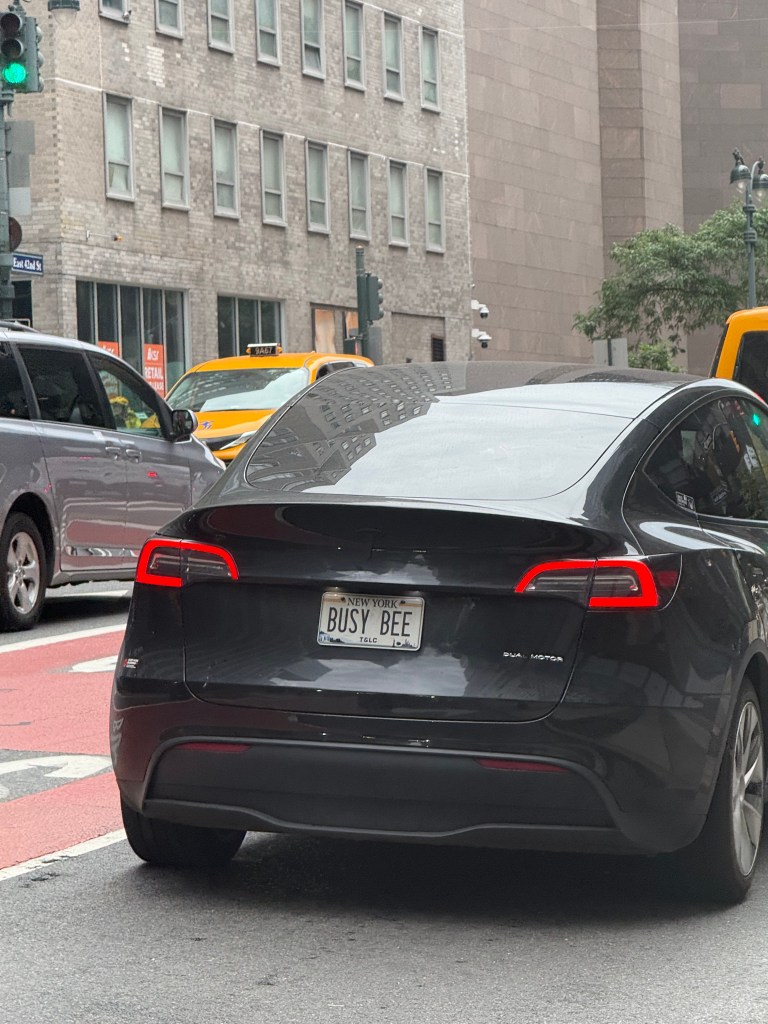 A black Tesla vehicle with a vanity license plate that reads 'BUSY BEE', parked on a city street with traffic and nearby buildings.
