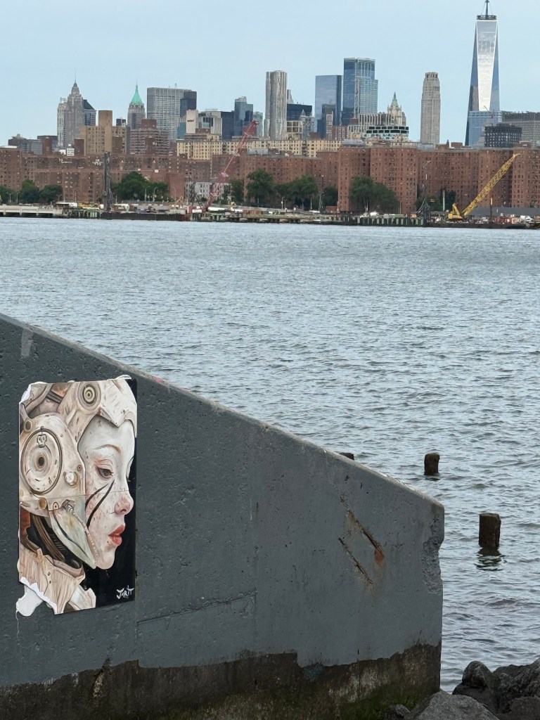 A detailed artwork of a robotic woman's face is mounted on a concrete structure by the waterfront, with the city skyline in the background.