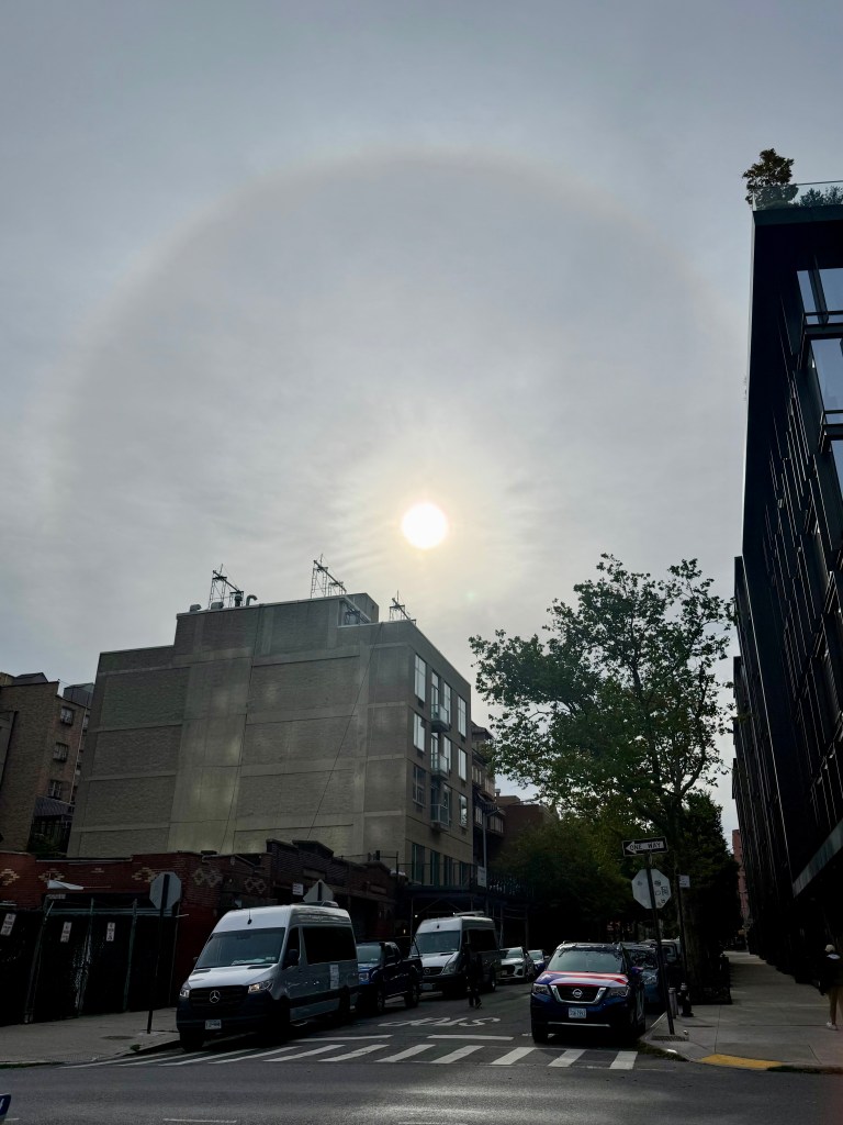 A view of the sun partially obscured by clouds, surrounded by a faint halo, with buildings and vehicles visible in the foreground.
