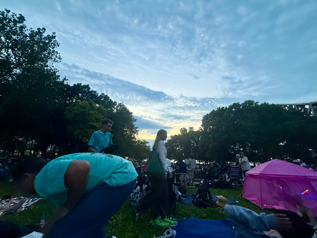 People enjoying an outdoor gathering at dusk, with trees and a colorful sky in the background.