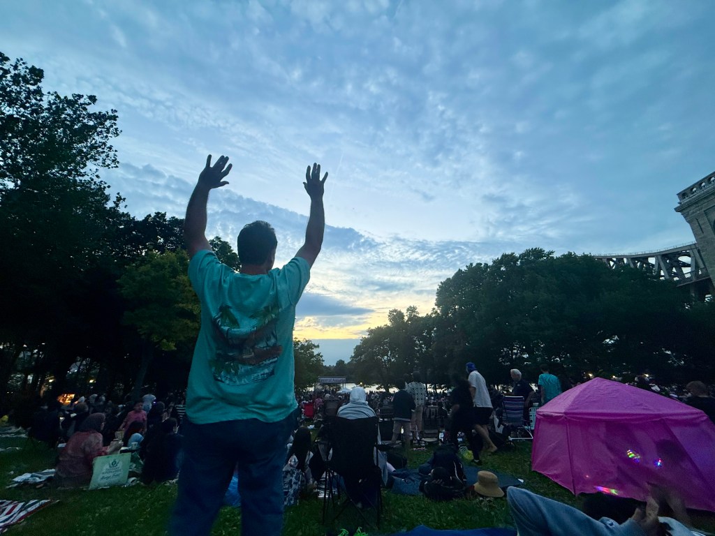 A crowd enjoying a sunset at an outdoor event, with a person in a turquoise shirt raising their arms in celebration.