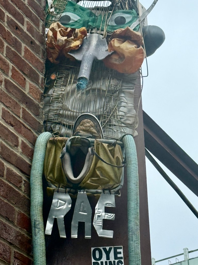 An abstract art installation mounted on a brick wall, featuring various materials such as a metallic structure, colored fabric, and eyes made from plastic. The piece includes the letters 'RAE' in a bold, metallic finish, alongside a sign that reads 'OYE PUNS'.