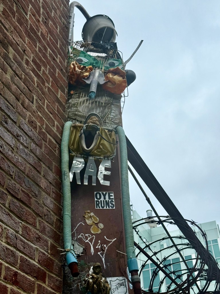 A colorful art installation on a brick wall featuring various materials, including metal, plastic, and clothing, resembling a whimsical character with green eyes and a mouth made of tubing, accompanied by graffiti and stickers.