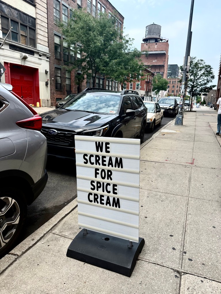 A sidewalk sign reading 'WE SCREAM FOR SPICE CREAM' with cars parked alongside a street lined with brick buildings and trees.