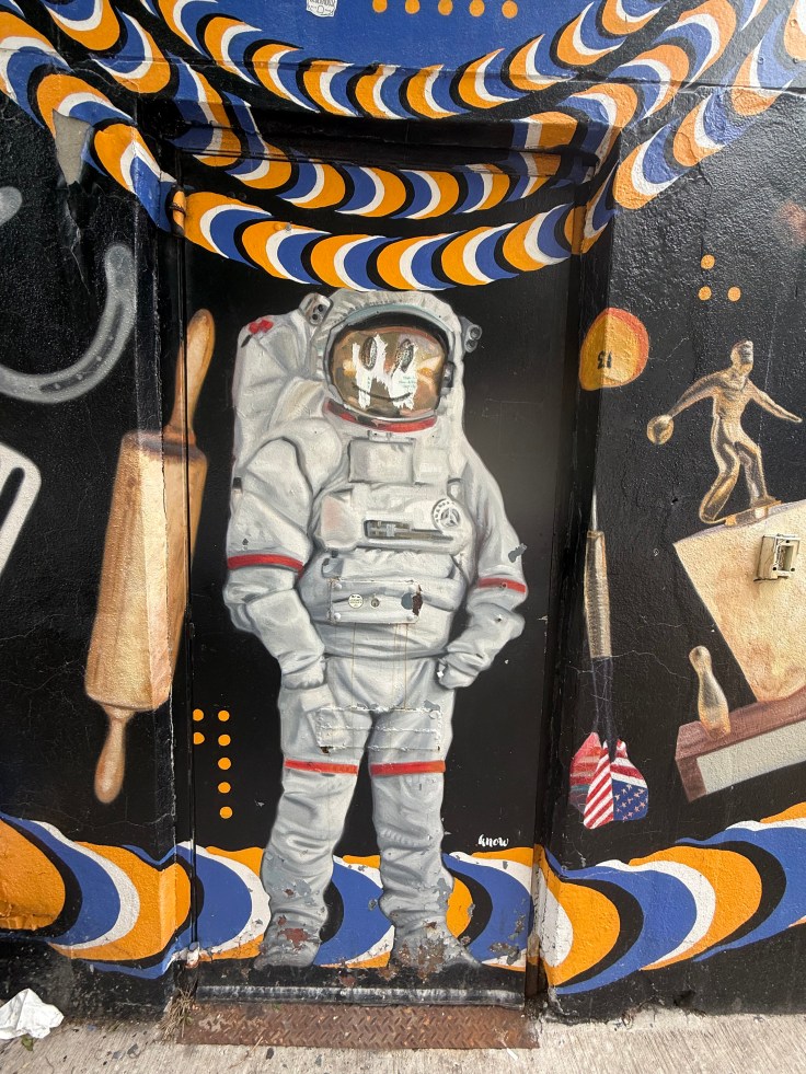 A mural featuring a cartoon-style astronaut in a spacesuit, standing in front of a door. The background includes vibrant abstract patterns in orange, blue, and white, along with various artistic elements like a rolling pin and a bowling figure.