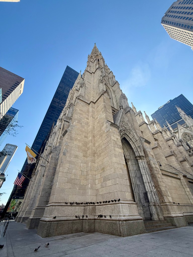 Low-angle view of a tall, ornate stone church surrounded by modern skyscrapers, under a clear blue sky.