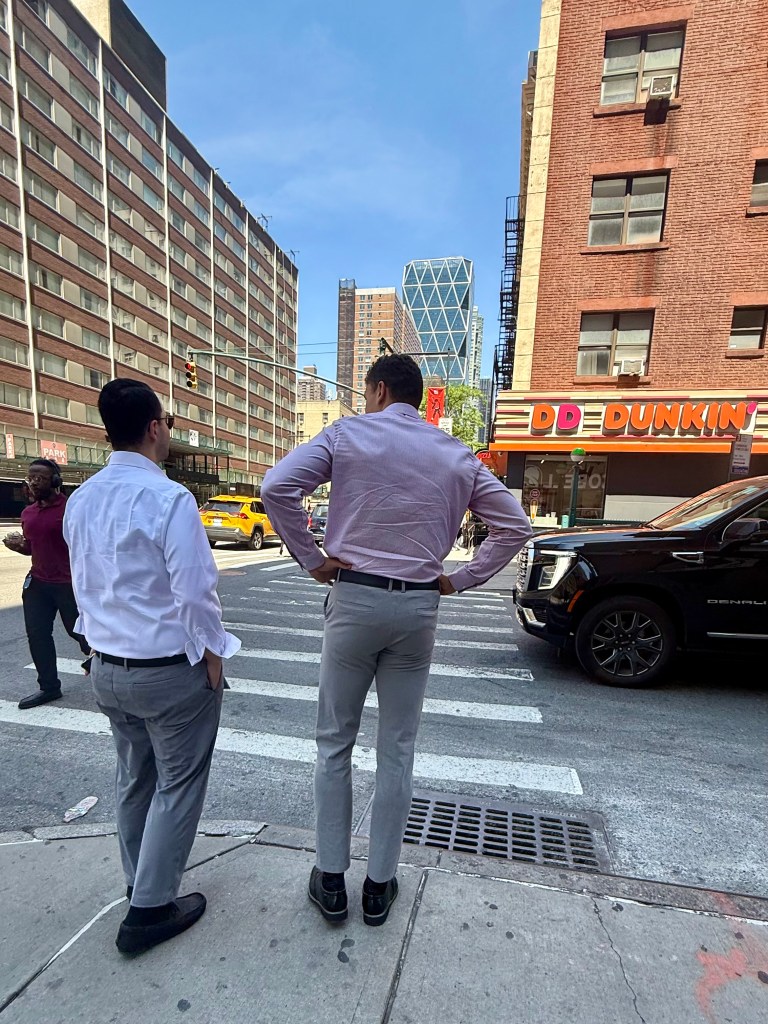Two men in business attire standing at a crosswalk in a city, with a Dunkin' Donuts shop and tall buildings in the background.