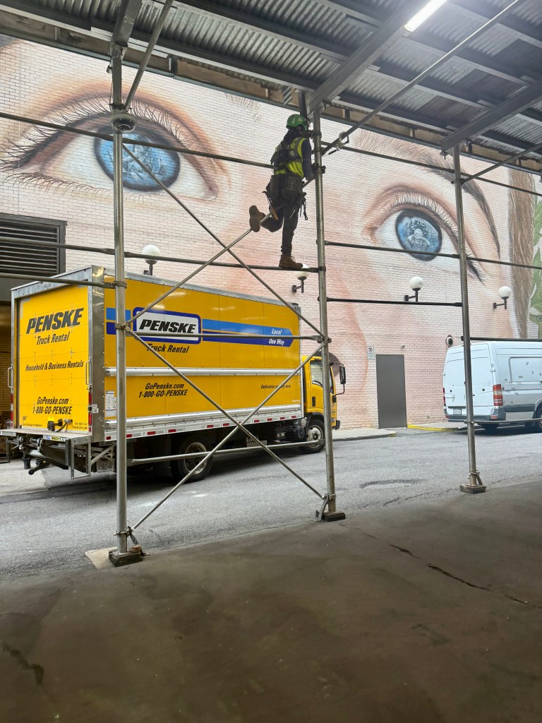 A worker in a safety harness climbs scaffolding next to a wall featuring a large mural of a blue eye. In the foreground, a yellow Penske moving truck is parked.