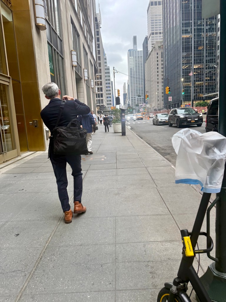 A man in a suit with a bag walks along a city sidewalk, photographing his surroundings, with tall buildings and traffic in the background.