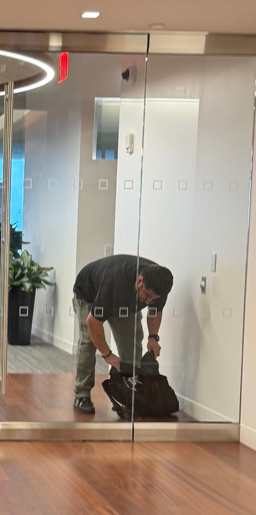 A person crouching down and looking into a black bag in an office corridor, with a mirrored glass wall and potted plants in the background.