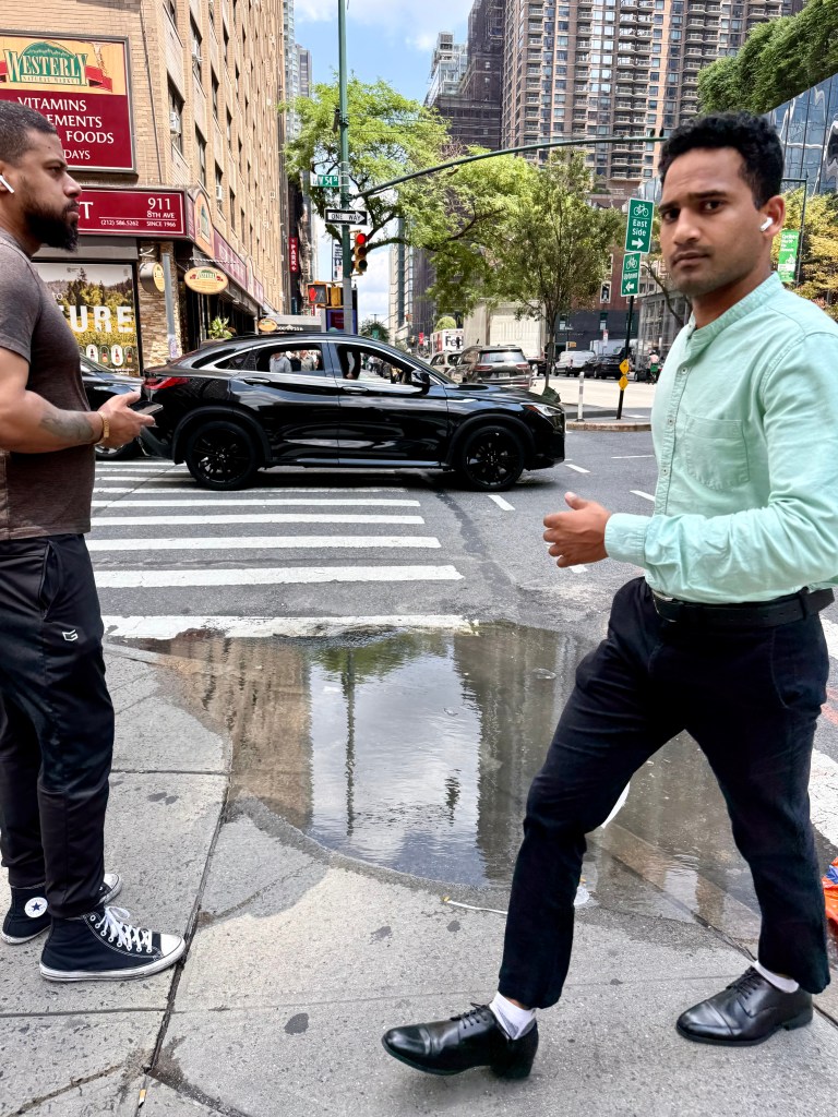 Two men crossing a street in an urban setting, one wearing a green shirt and the other in a dark t-shirt, with a black car driving past and a puddle on the sidewalk.