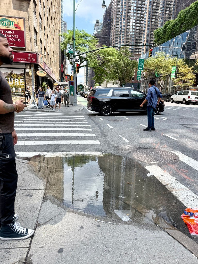 A busy street corner in an urban environment featuring a puddle reflecting buildings and traffic lights, with pedestrians crossing the street and vehicles on the road.