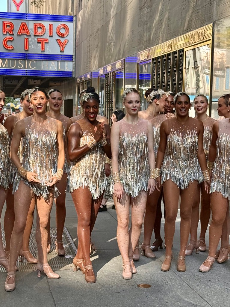 A group of dancers in shimmering costumes with fringes, standing in front of the Radio City Music Hall sign. They are smiling and posing for the camera in a lively outdoor setting.