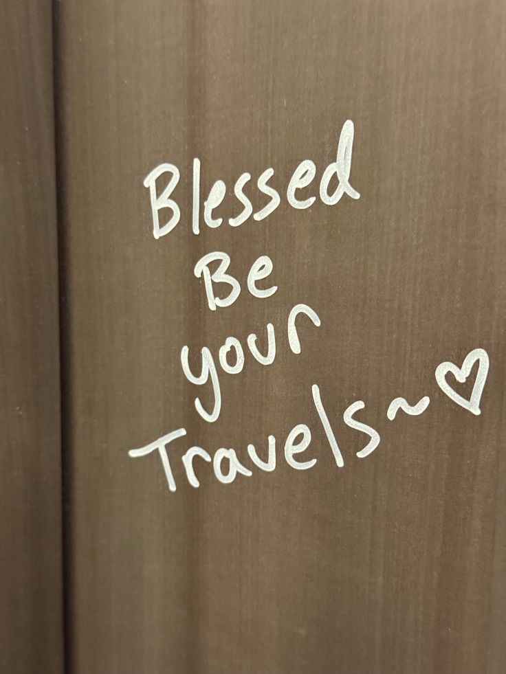 A wooden surface with handwritten text that says 'Blessed Be your Travels~' followed by a heart symbol.