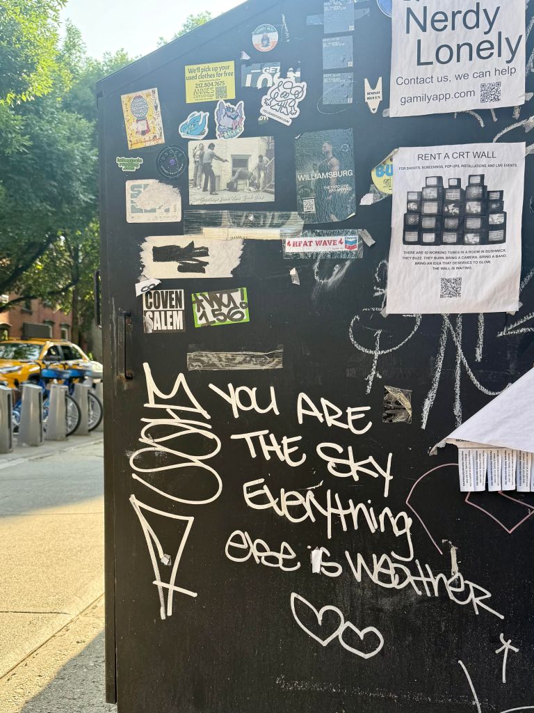 A black utility box covered with various stickers and flyers, displaying graffiti text that reads 'YOU ARE THE SKY everything else is WEATHER.' The background includes trees and bicycles parked nearby.