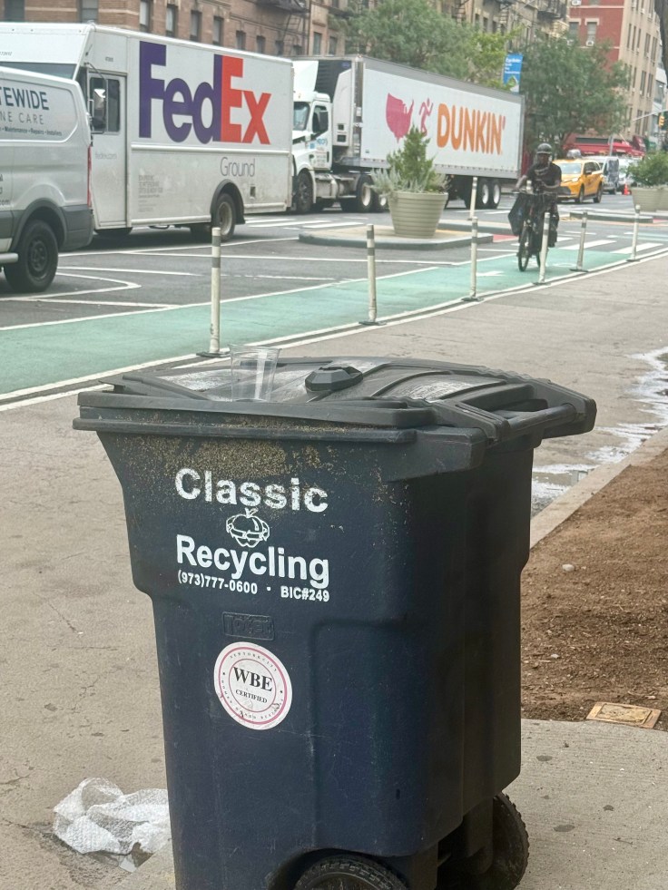 A blue recycling bin labeled 'Classic Recycling' on a city street, with delivery trucks visible in the background.